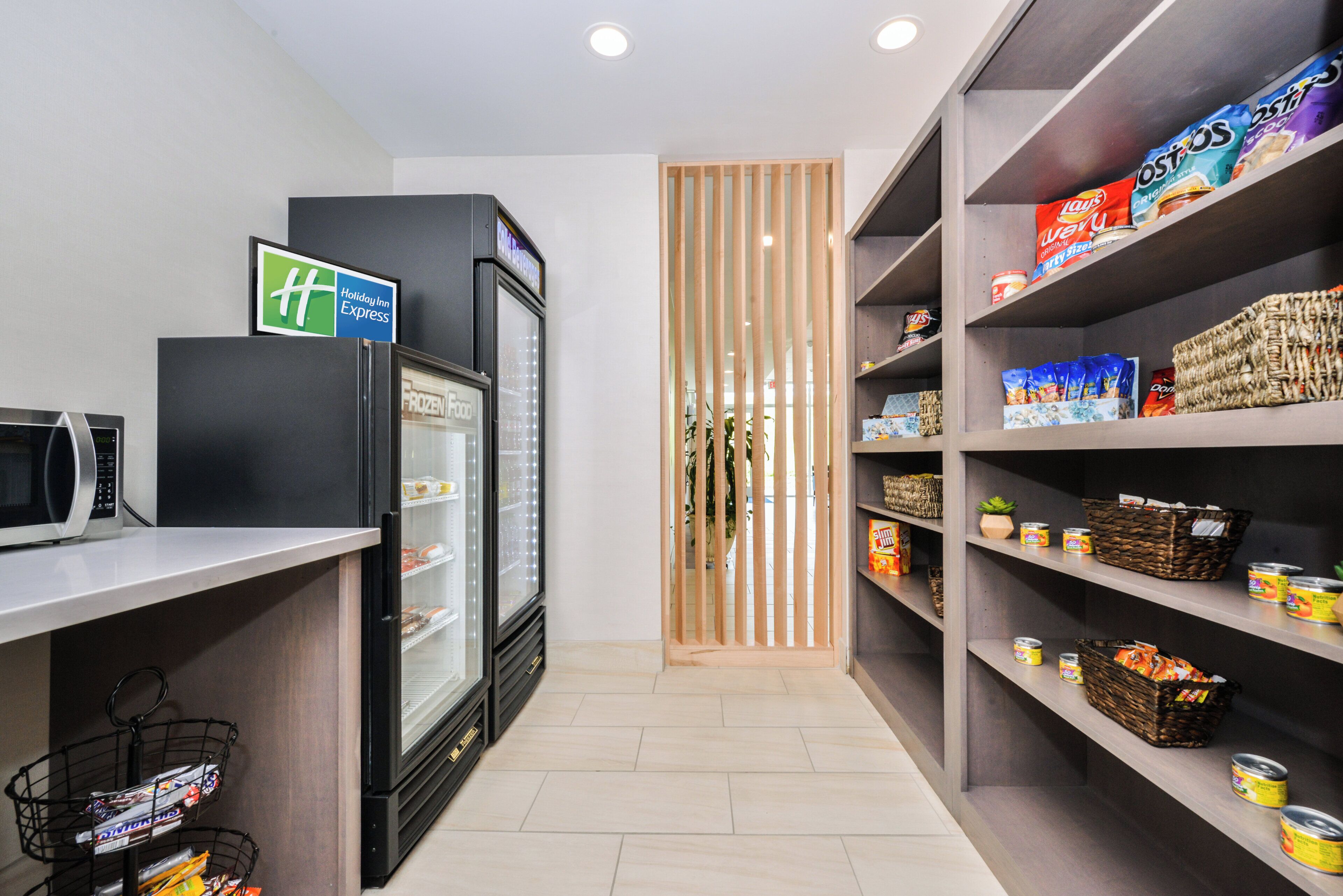 A hotel kitchenette with a microwave, refrigerator, and shelves stocked with snacks and beverages.