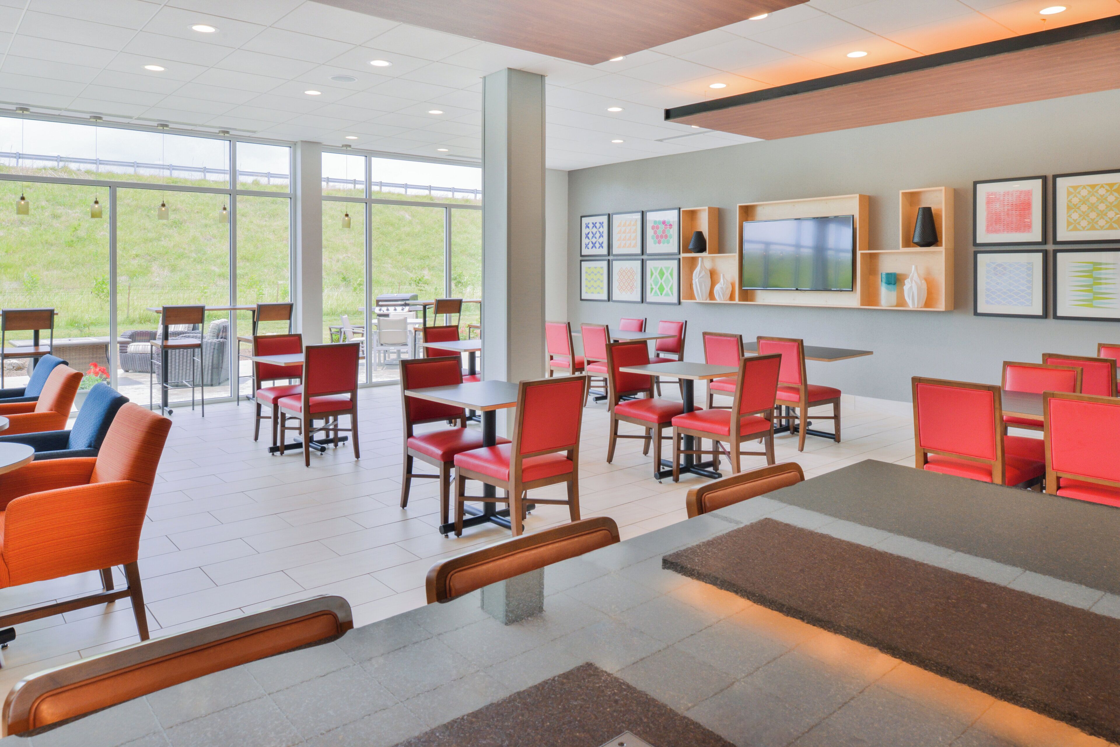 A modern dining area with red and orange chairs, a flat-screen TV on the wall, and a view of a grassy field through large windows.