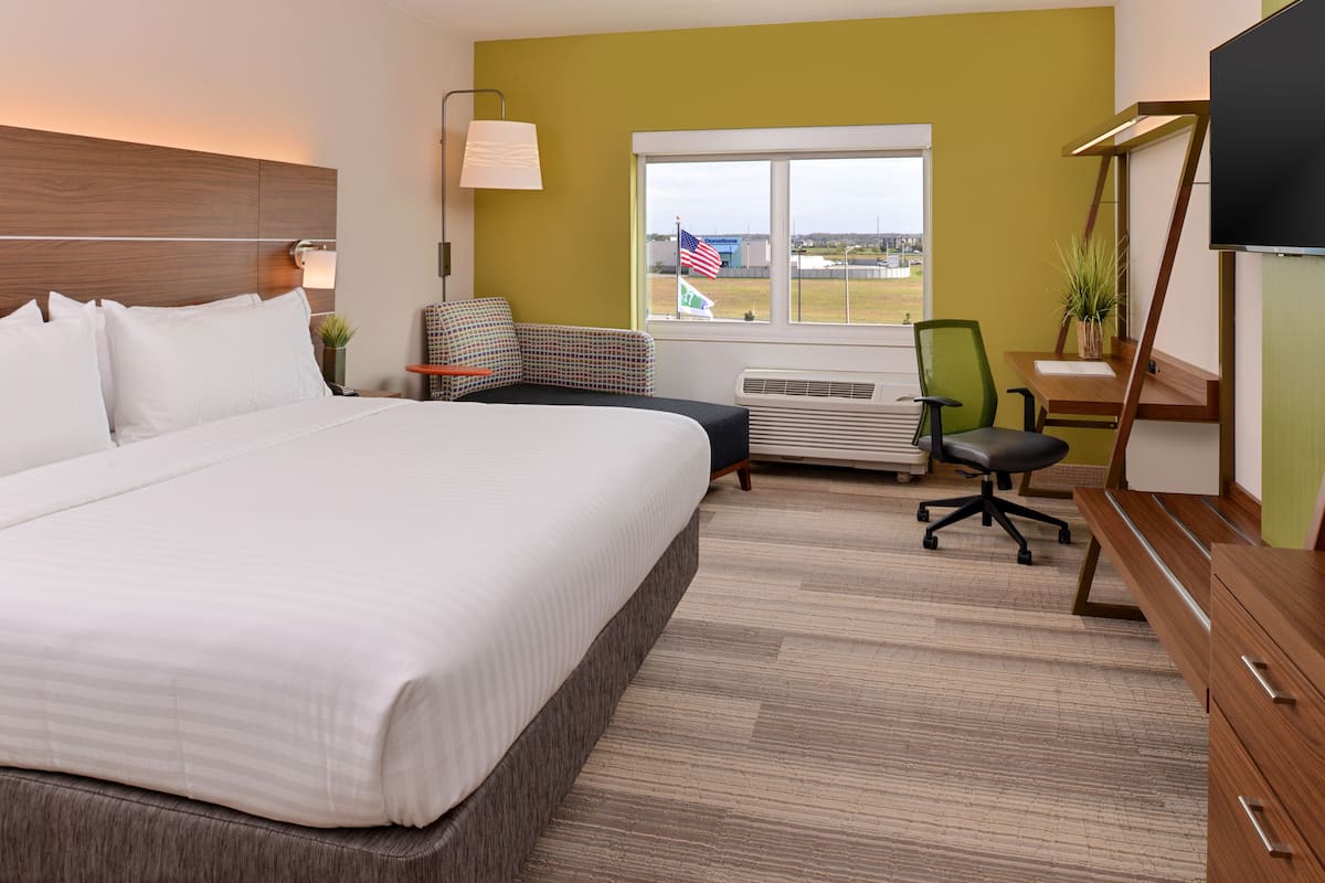 A hotel room with a large bed, a desk with a chair, a TV, and a window with an American flag view.