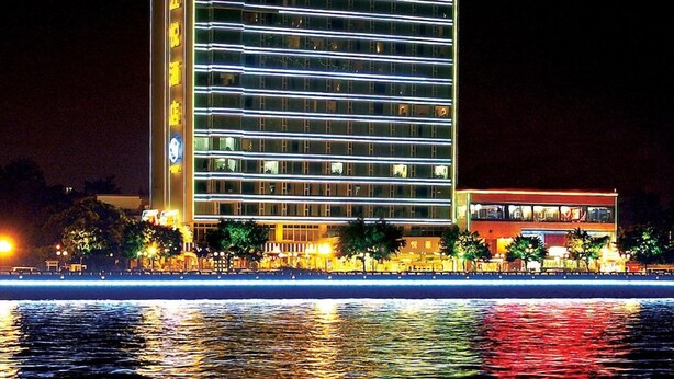 Un hotel de varios pisos con señalización iluminada, un reloj a la vista y un reflejo en el agua frente al edificio.