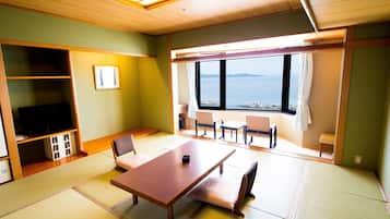 A room with tatami flooring, a wooden table, chairs, a television, and a view of the sea.