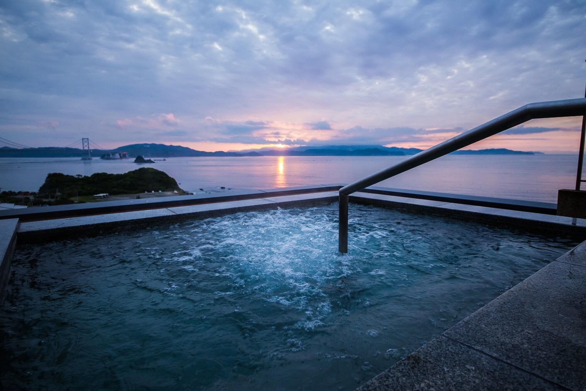 A hot tub with a view of a sunset over water, a bridge in the distance, and a railing.