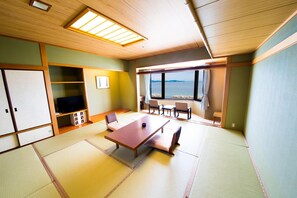 A traditional Japanese-style room with tatami flooring, a low table, chairs, a television, and a large window offering a view of the sea. - Naruto Grand Hotel (Naruto)