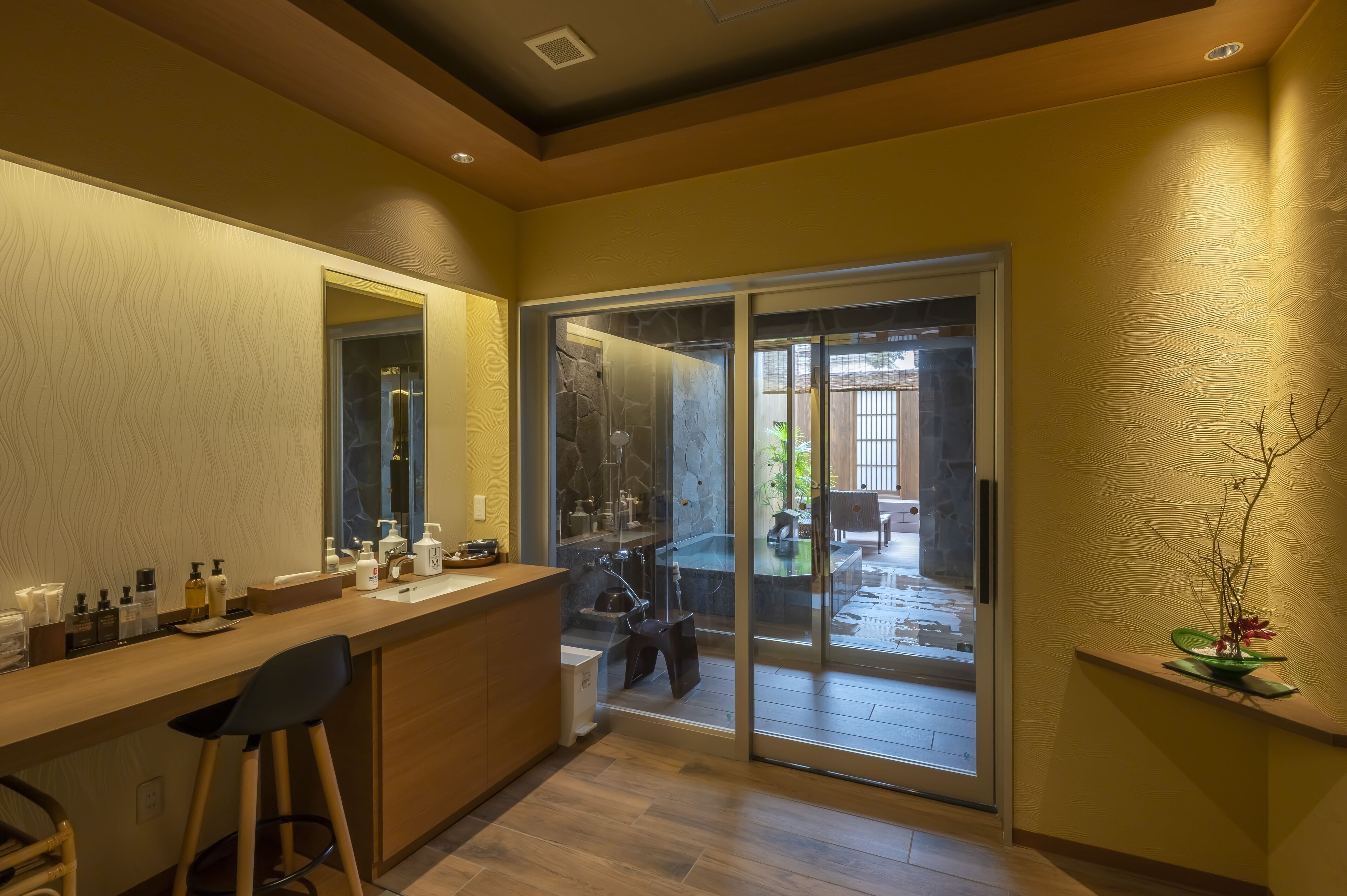 A modern bathroom with a glass shower, a vanity with toiletries, and a view of the outdoor patio.