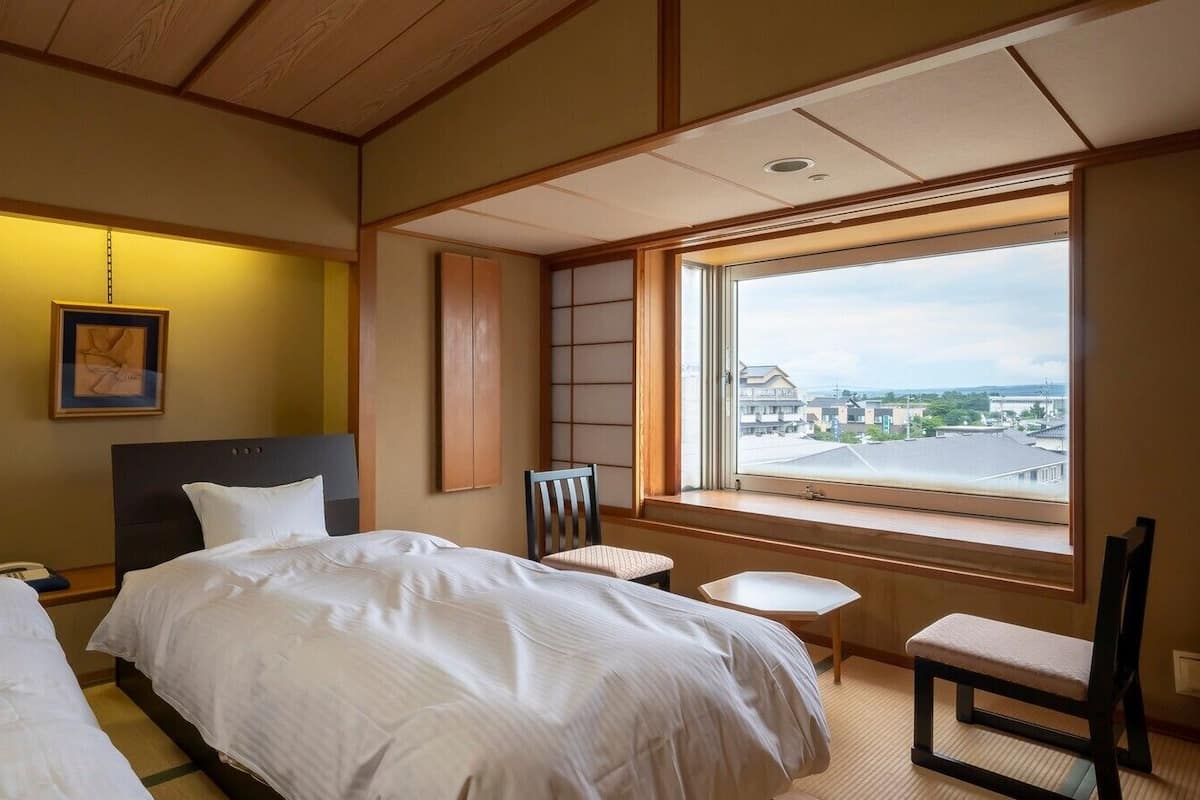 A hotel room with two beds, a small table, a chair, a window with a city view, and a framed picture on the wall.