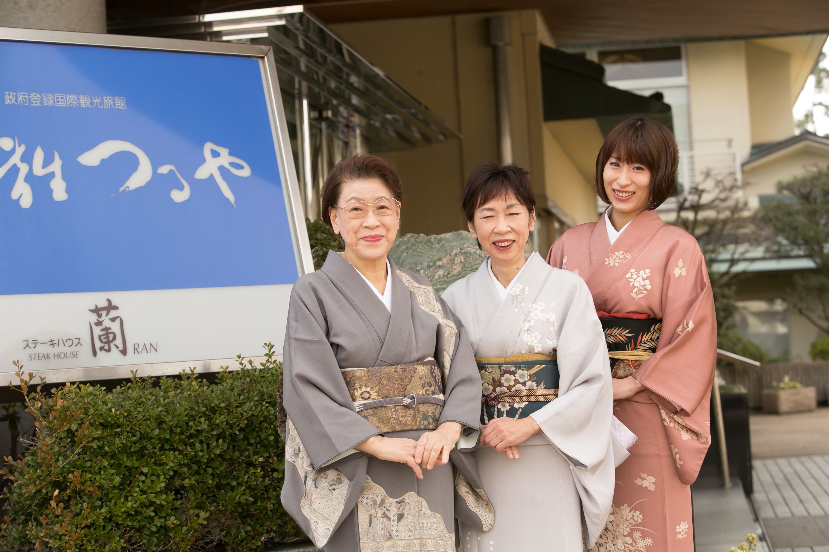 Three people in traditional Japanese attire standing in front of a building with a sign that reads "STEAK HOUSE."