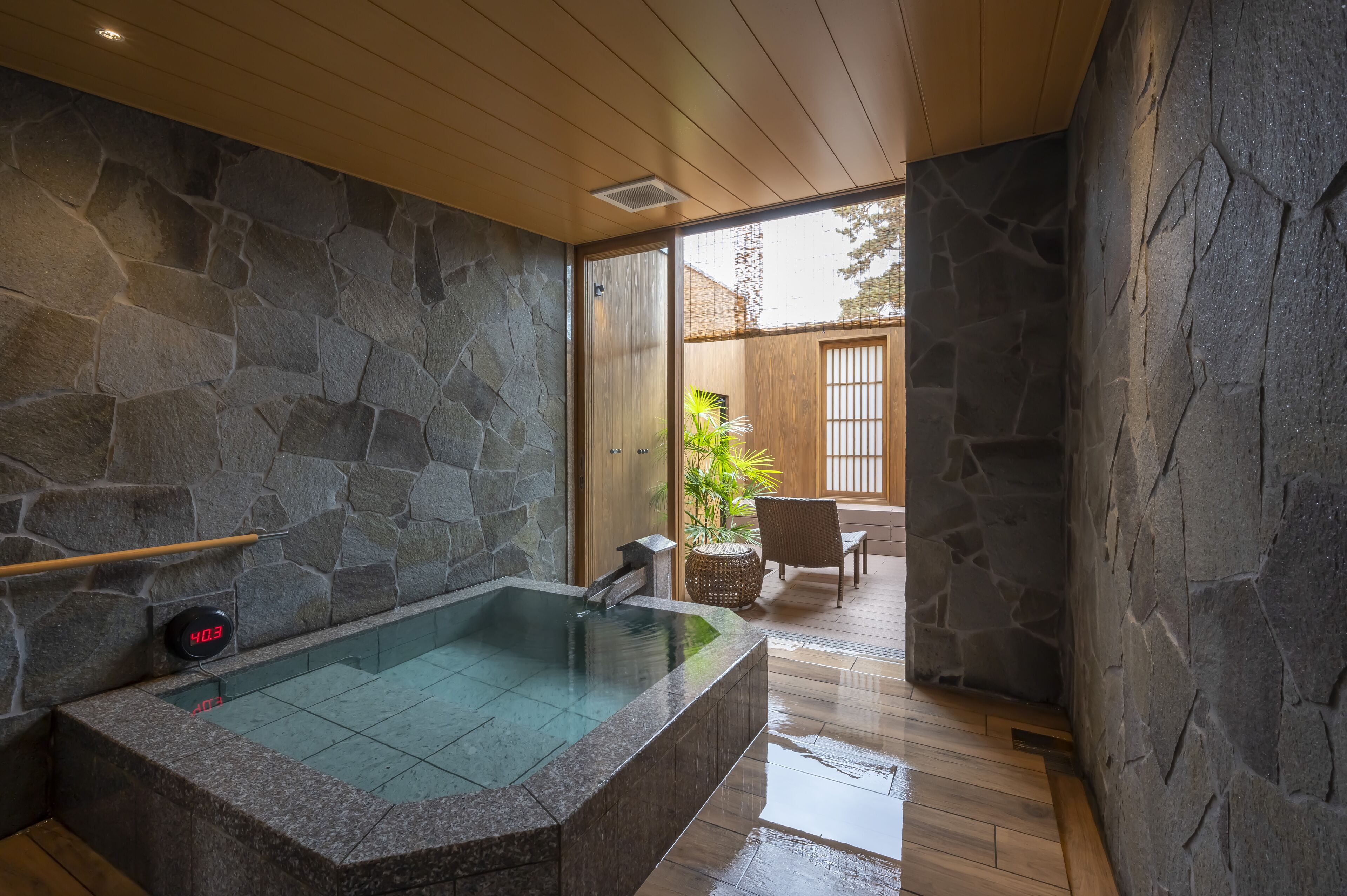 A stone-walled bathroom with a large bathtub, wooden flooring, and a view of a garden through a sliding door.