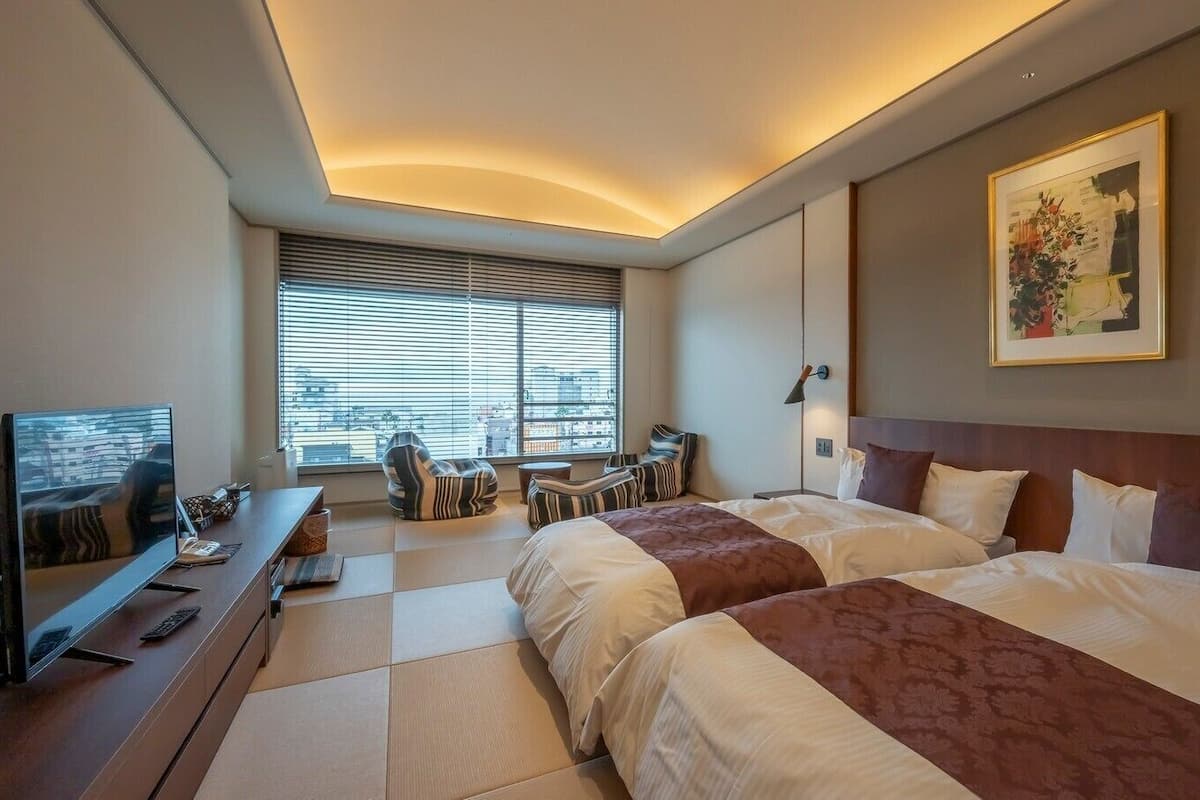 Habitación de hotel con dos camas, televisión, escritorio y ventana con persianas.