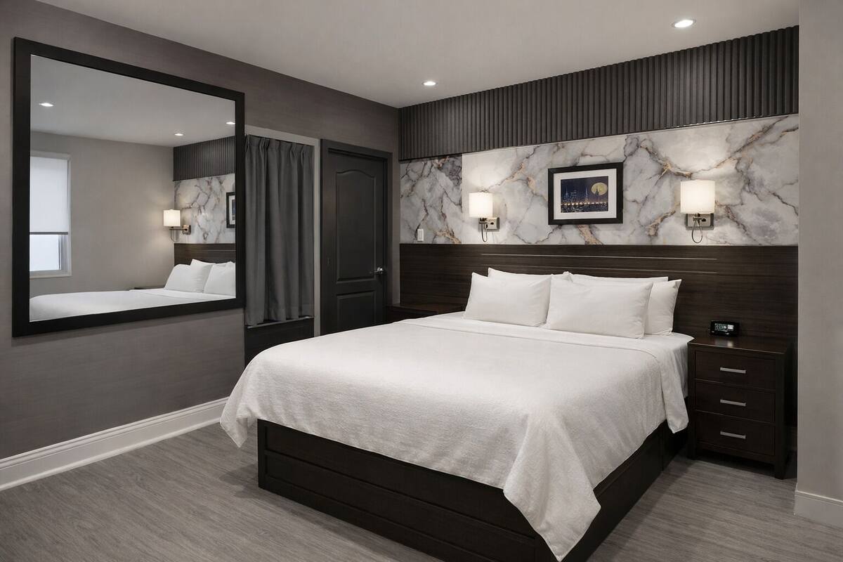 Quarto moderno com uma cama grande, cabeceira de madeira escura, mesa de cabeceira com abajur, quadro na parede e espelho sobre a cama.
