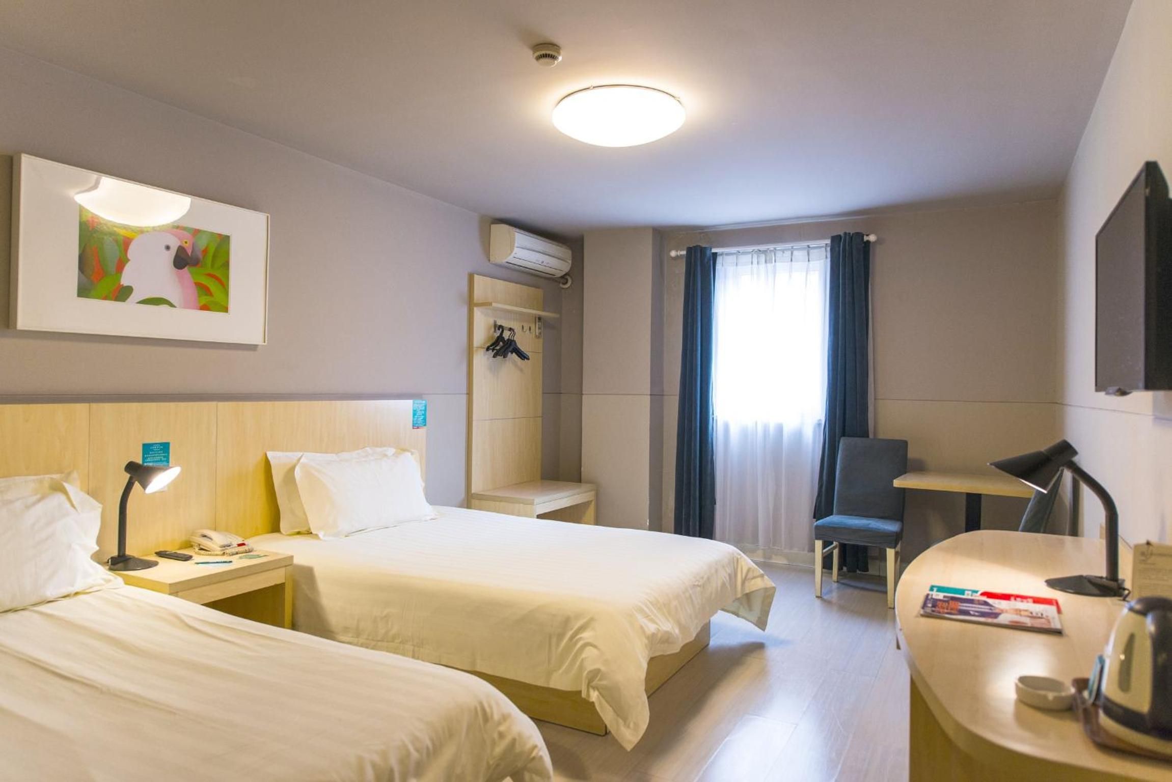 A hotel room with two beds, a desk, a chair, a small table, a wall-mounted air conditioner, a framed picture, and a window with curtains.