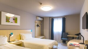 Camera d'albergo con due letti, una scrivania, una sedia, un tavolino, un condizionatore d'aria a parete, un quadro incorniciato e una finestra con tende.