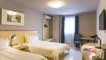 A hotel room with two beds, a desk, a chair, a small table, a wall-mounted air conditioner, a framed picture, and a window with curtains.