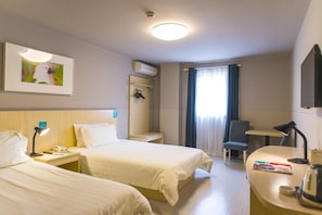 Quarto de hotel com duas camas, uma escrivaninha, uma cadeira, uma mesinha, ar condicionado fixado na parede, um quadro emoldurado e uma janela com cortinas.