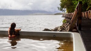 Iemand zit in een jacuzzi aan een meer, met een houten terras en een rotsachtige oever.