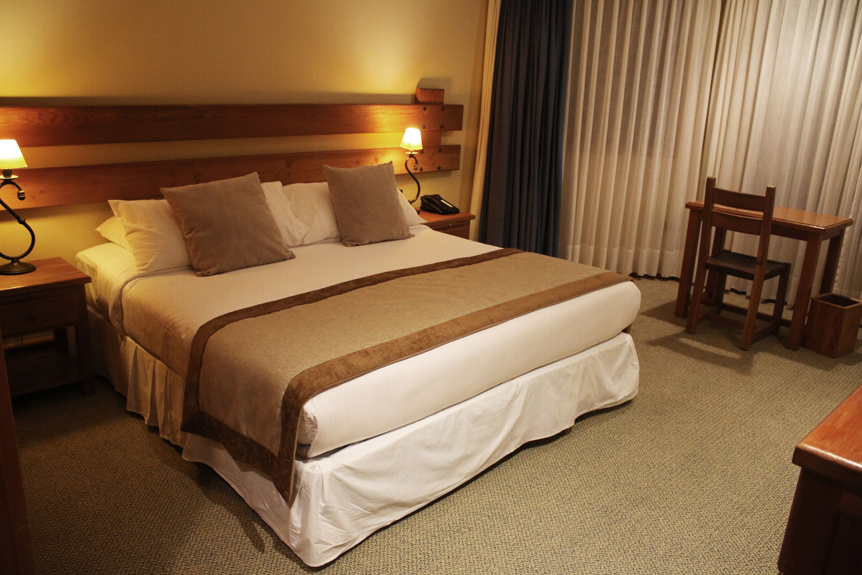 Hotelkamer met een groot bed, twee nachtkastjes met lampen, een houten hoofdeinde, een stoel en een gordijn.