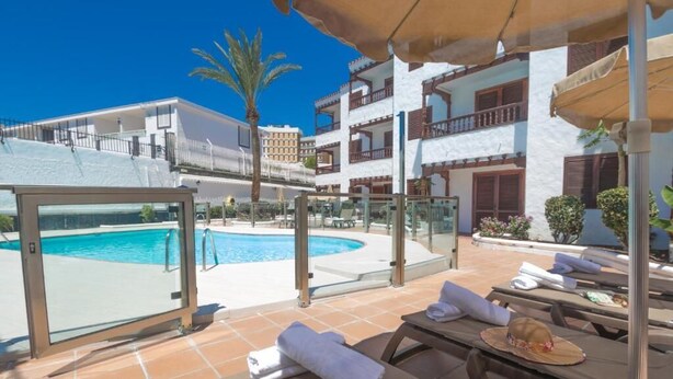 A poolside area with sun loungers, a glass-enclosed pool, and a view of a multi-story building with balconies. - Alsol Las Orquideas (San Bartolomé de Tirajana)
