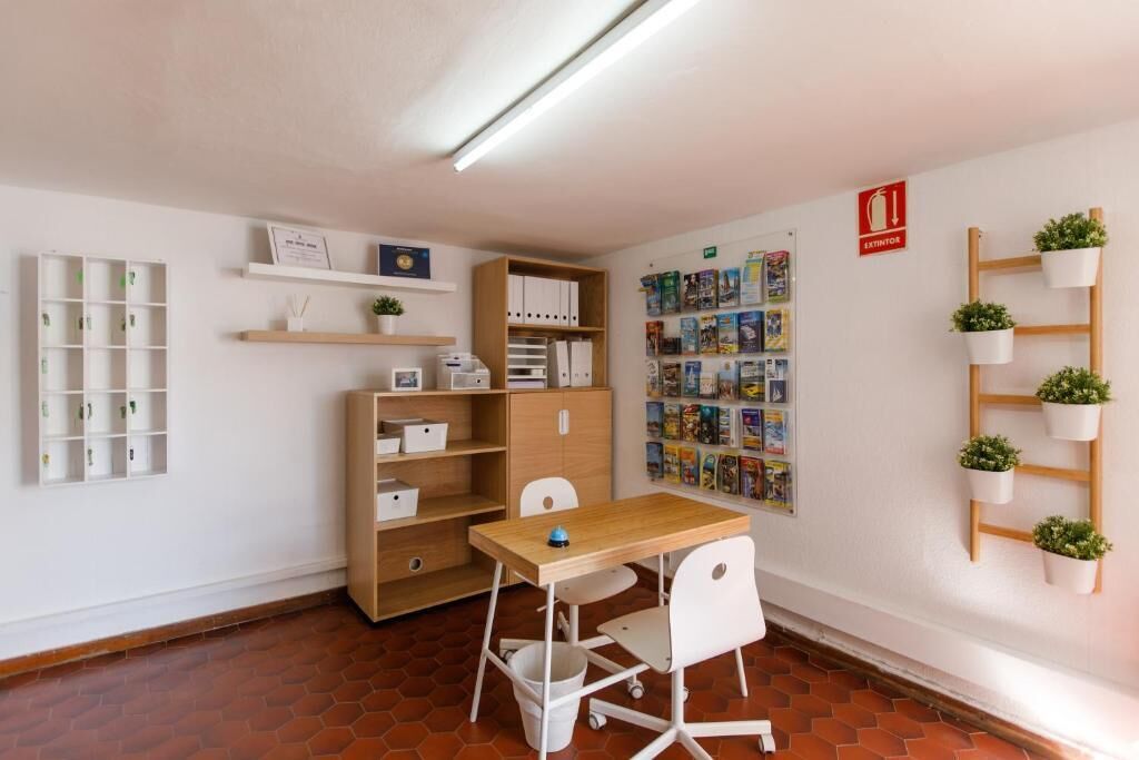 A room with a wooden desk, white chairs, a bulletin board with magazines, shelves with binders, and a fire exit sign.