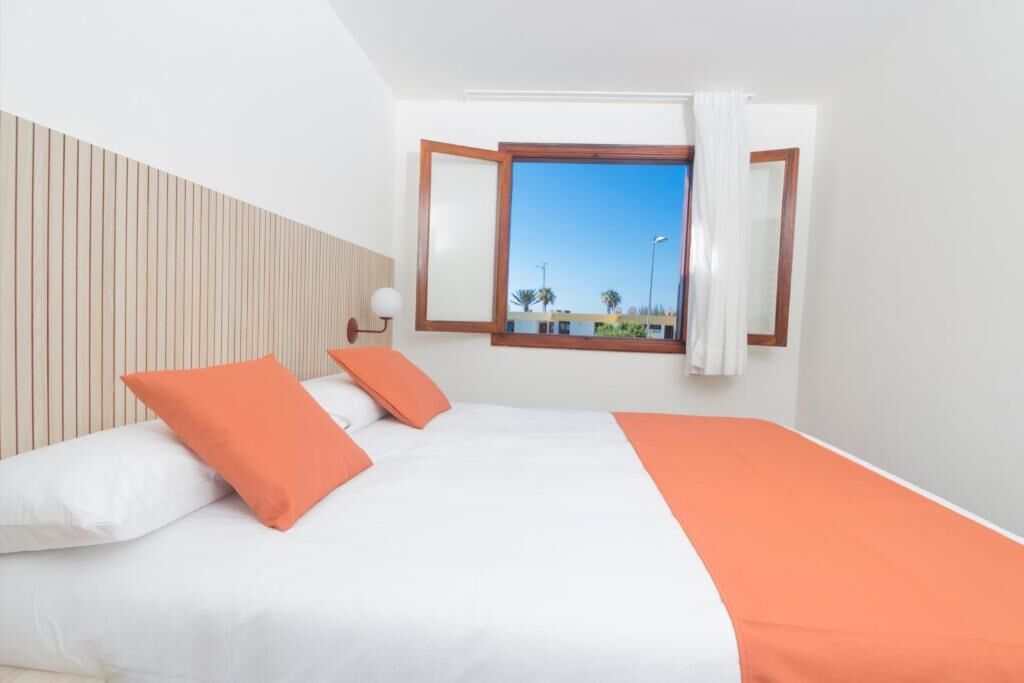 A hotel room with a bed, white and orange bedding, a wooden headboard, a window with curtains, and a view of palm trees and buildings.