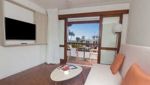 A hotel room with a TV, a sofa, a small table, and a balcony with a view of palm trees and a building. - Alsol Las Orquideas (San Bartolomé de Tirajana)