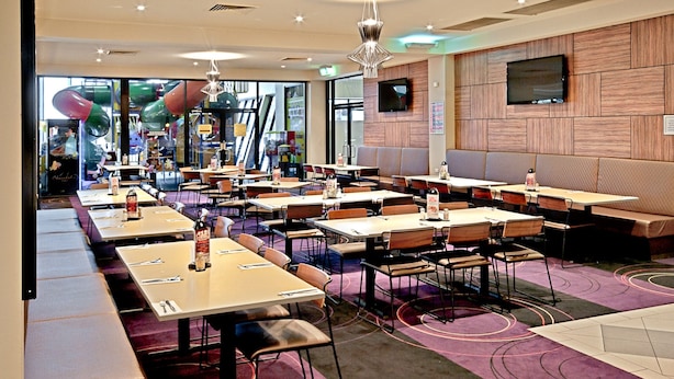 Un restaurante moderno con varias mesas y cabinas, un amplio ventanal con vistas a un parque infantil con toboganes, y una televisión de pantalla plana montada en la pared.