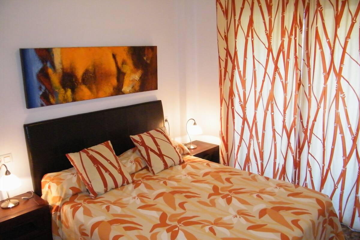 A bed with a floral bedspread, two pillows, a nightstand with a lamp, and a wall-mounted painting.