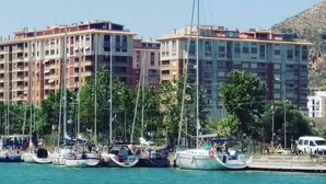 Uma marina com vários barcos atracados, uma carrinha branca na margem, e um edifício de vários andares ao fundo.