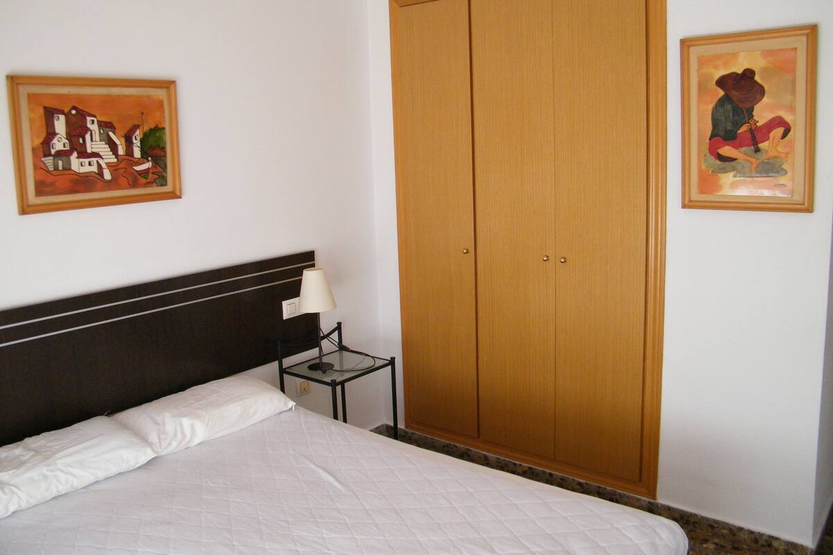 A bedroom with a bed, a nightstand, a wooden wardrobe, and two framed artworks on the walls.