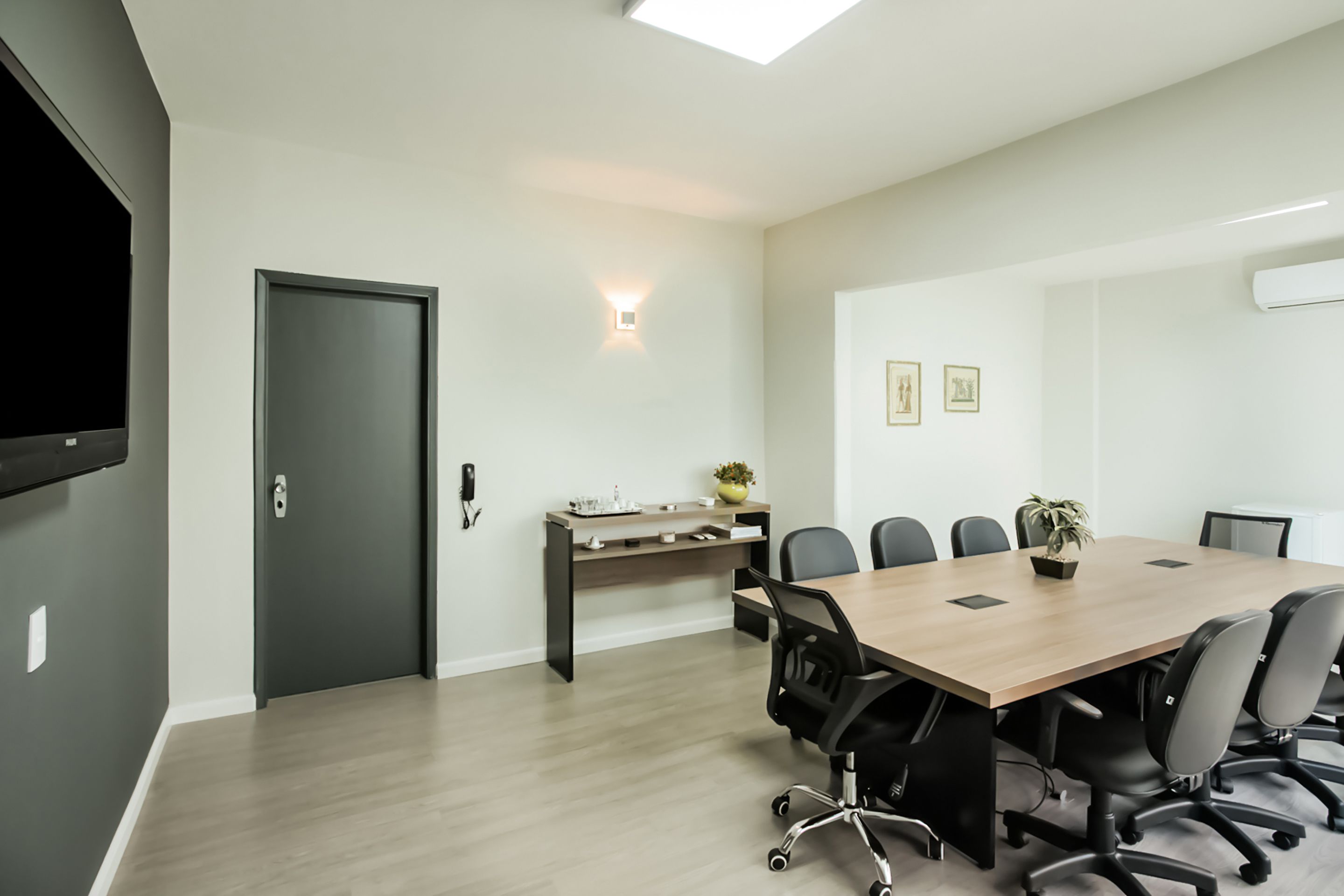 A conference room with a long table, chairs, a desk with a plant, and a wall-mounted TV.