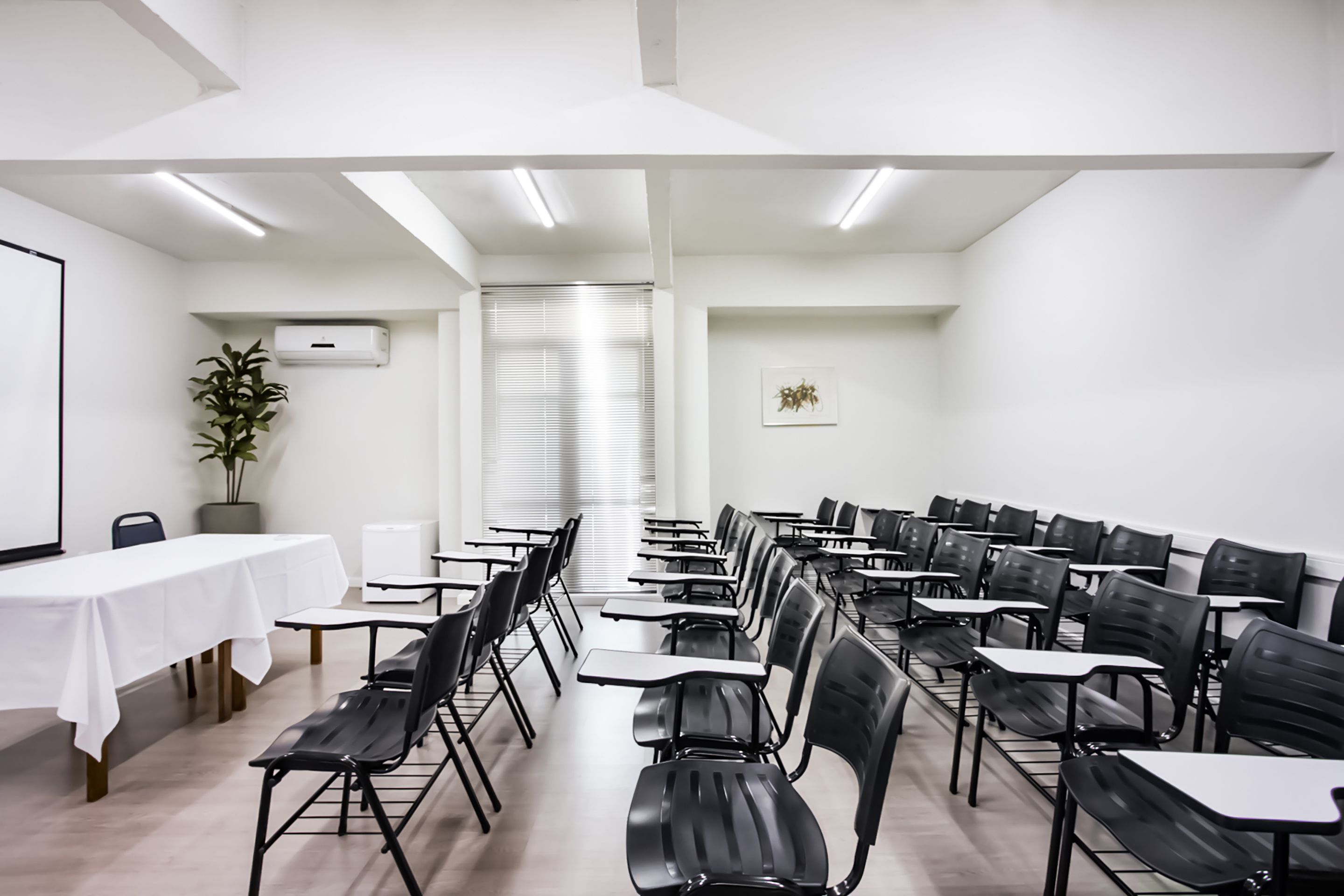 Sala de conferência com fileiras de cadeiras e mesas pretas, paredes brancas e uma tela de projetor.