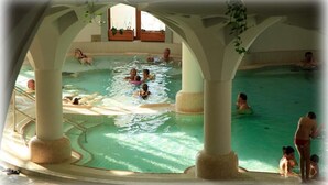 A large indoor swimming pool with multiple people swimming and playing, surrounded by white columns and a tiled floor. - Grand Hotel Glorius (Mako)