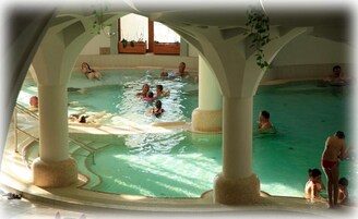 Una amplia piscina interior con varias personas nadando y jugando, rodeada de columnas blancas y un suelo de baldosas.