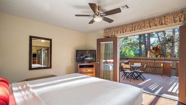 A bedroom with a bed, a ceiling fan, a TV, a dresser, and a sliding glass door leading to an outdoor deck with a table and chairs. - Roundhouse Resort (Pinetop)