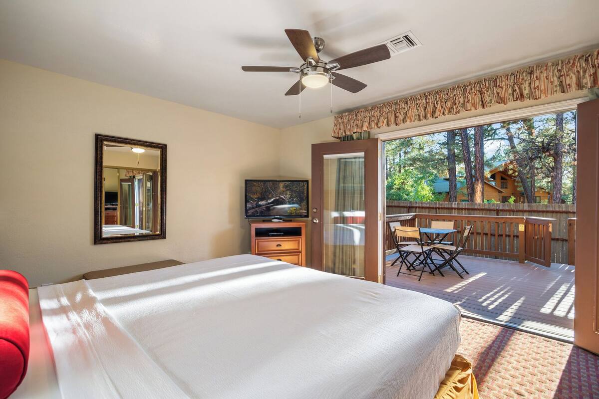 A bedroom with a bed, a ceiling fan, a TV, a dresser, and a sliding glass door leading to an outdoor deck with a table and chairs.