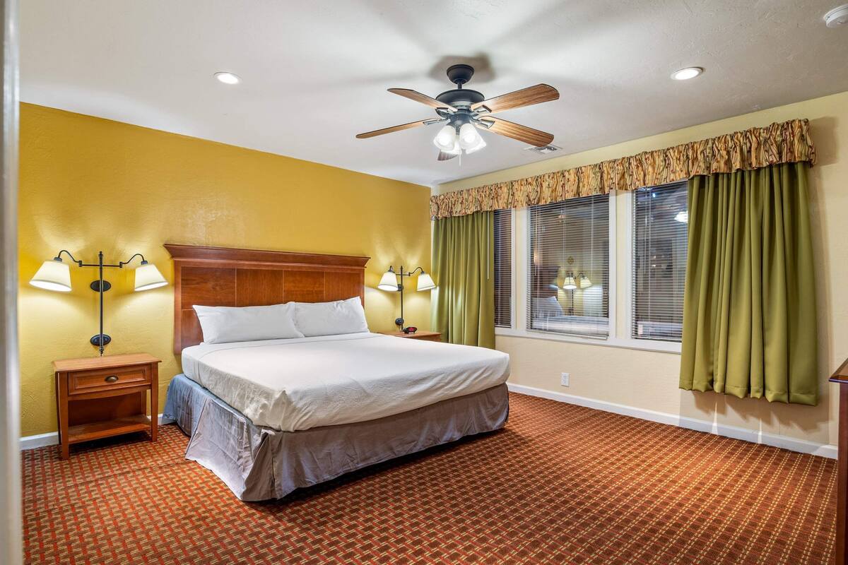 A hotel room with a bed, two bedside lamps, a ceiling fan, and a window with curtains.