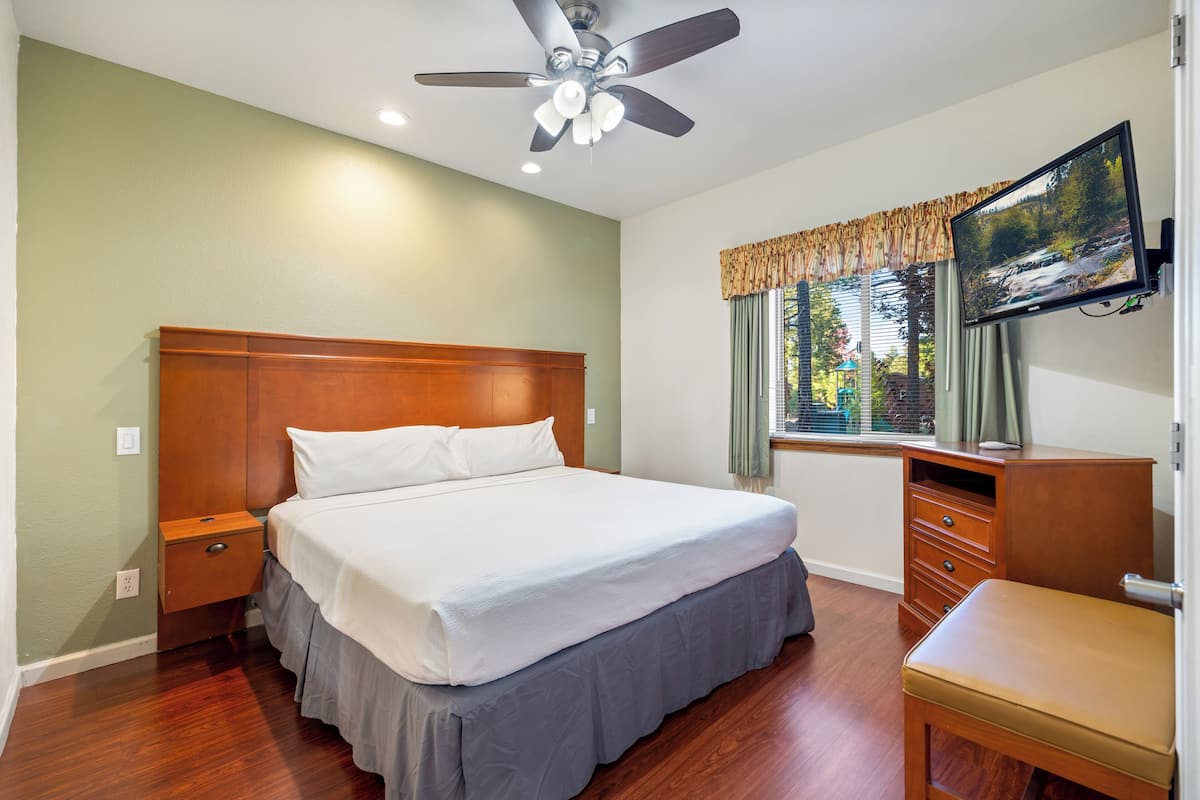 A neatly arranged bedroom with a bed, bedside tables, a desk with a chair, a ceiling fan, and a TV mounted on the wall.