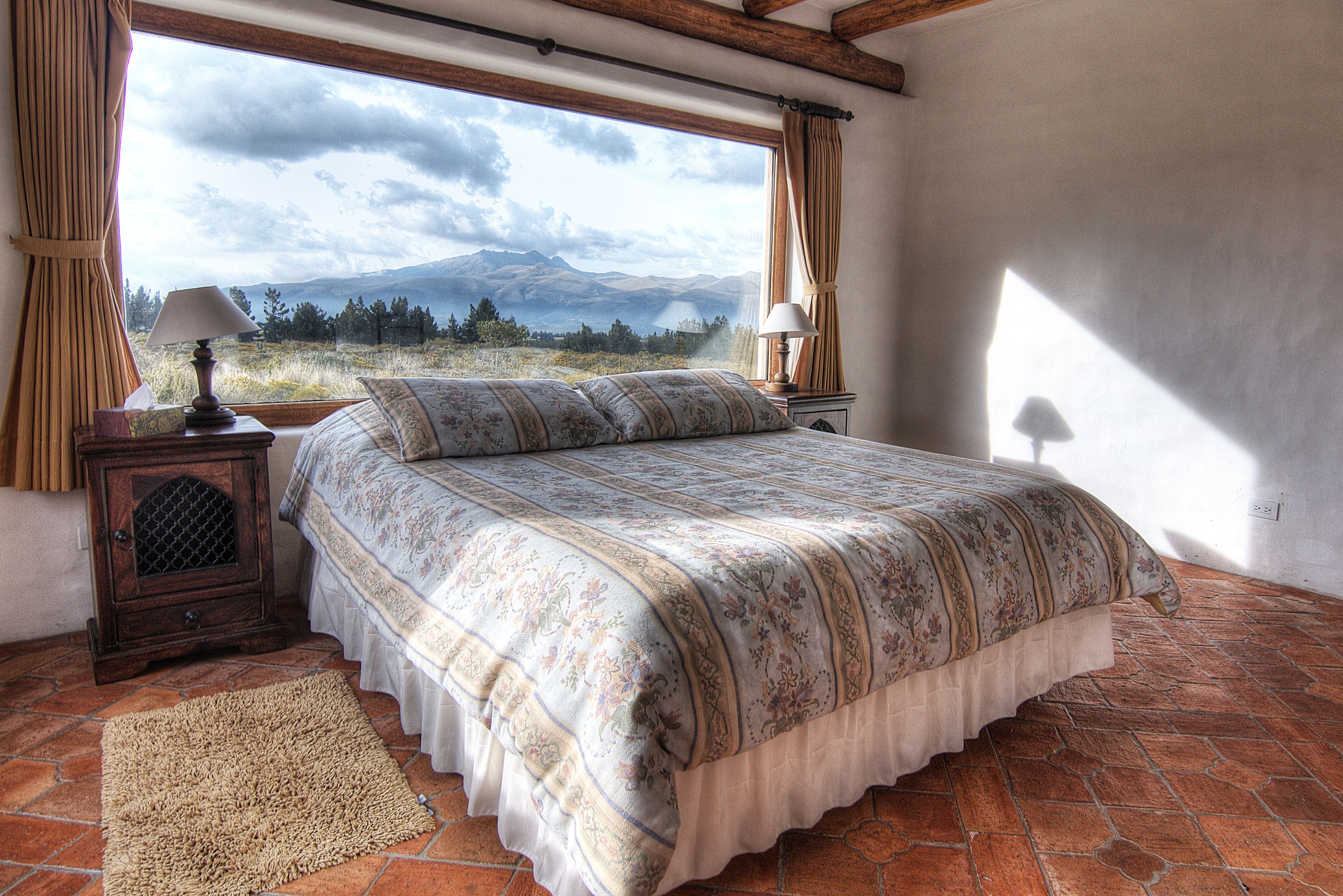Une chambre à coucher avec un grand lit, une table de chevet, une lampe et une vue sur les montagnes par la fenêtre.
