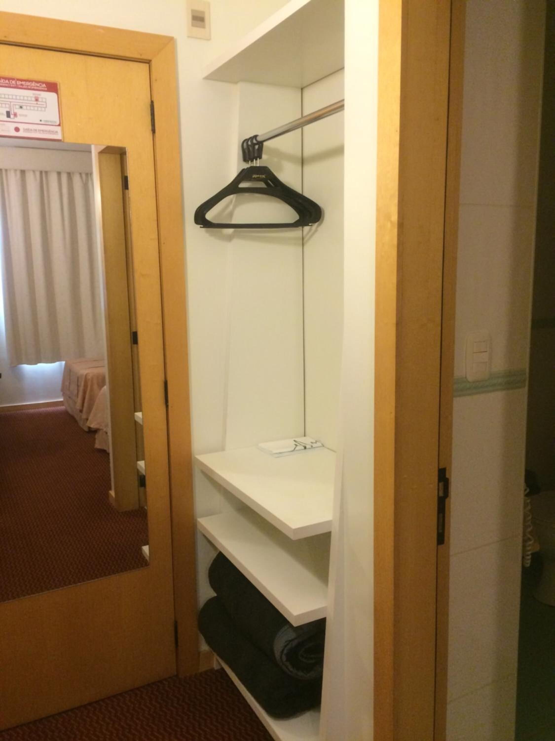 Una habitación de hotel con puerta de madera, espejo, estante blanco con perchero negro y cama con cubrecama blanco.