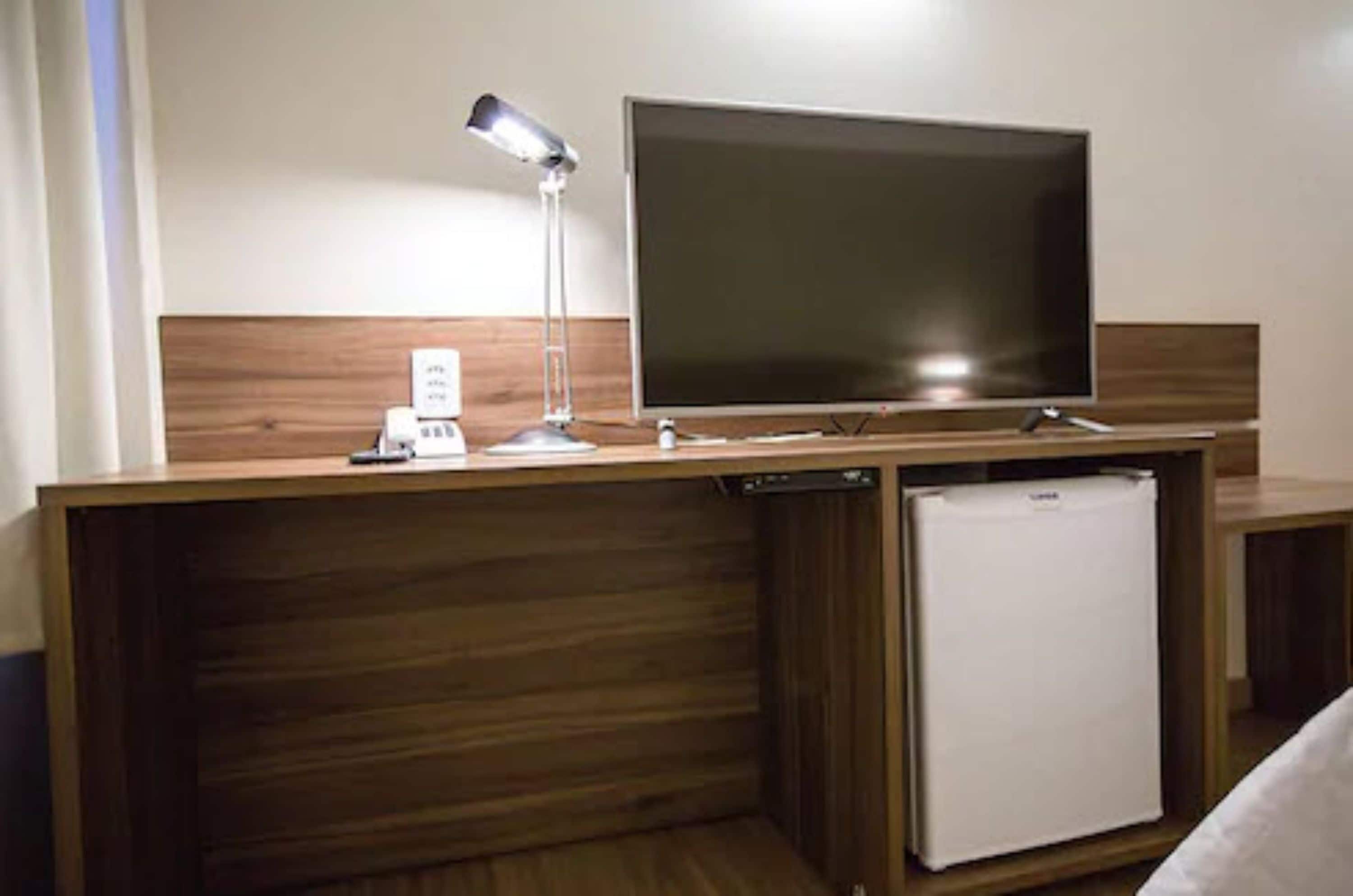 Une chambre d’hôtel dotée d’une tête de lit en bois, d’un téléviseur à écran plat, d’un bureau avec une lampe, d’un mini-réfrigérateur et d’une prise de courant.