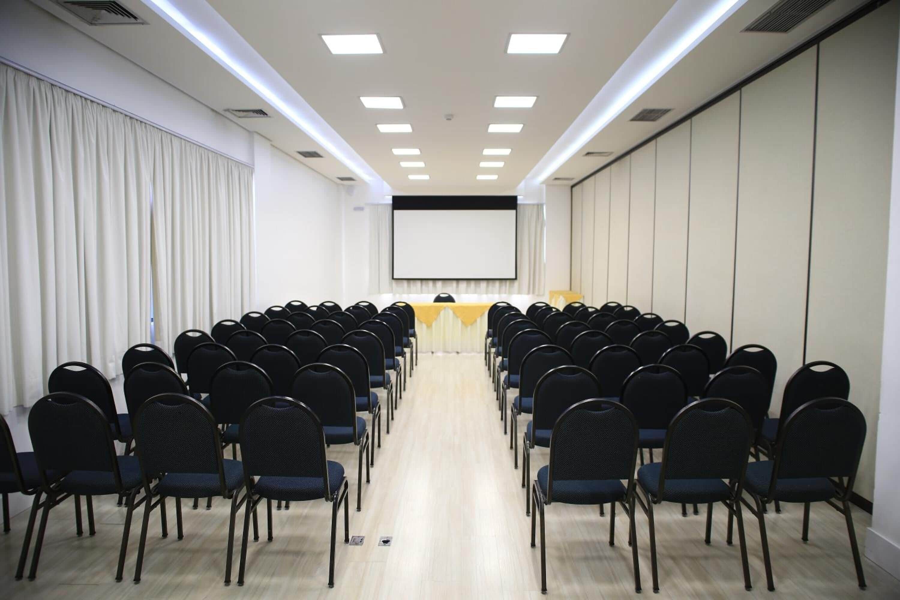 A conference room with rows of chairs facing a stage with a screen, equipped with ceiling lights and a curtain backdrop.