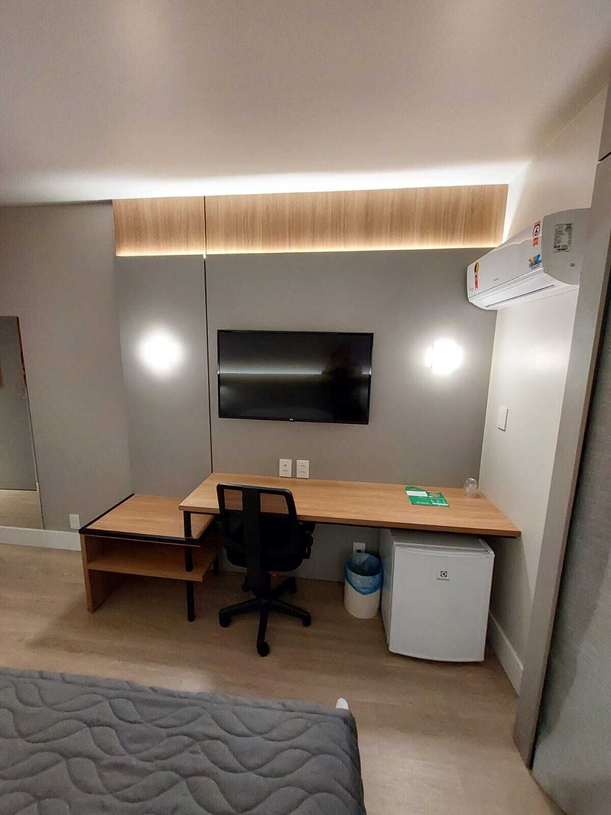 Habitación de hotel pequeña con una cama individual, un escritorio con silla, un televisor montado en la pared y un refrigerador pequeño.