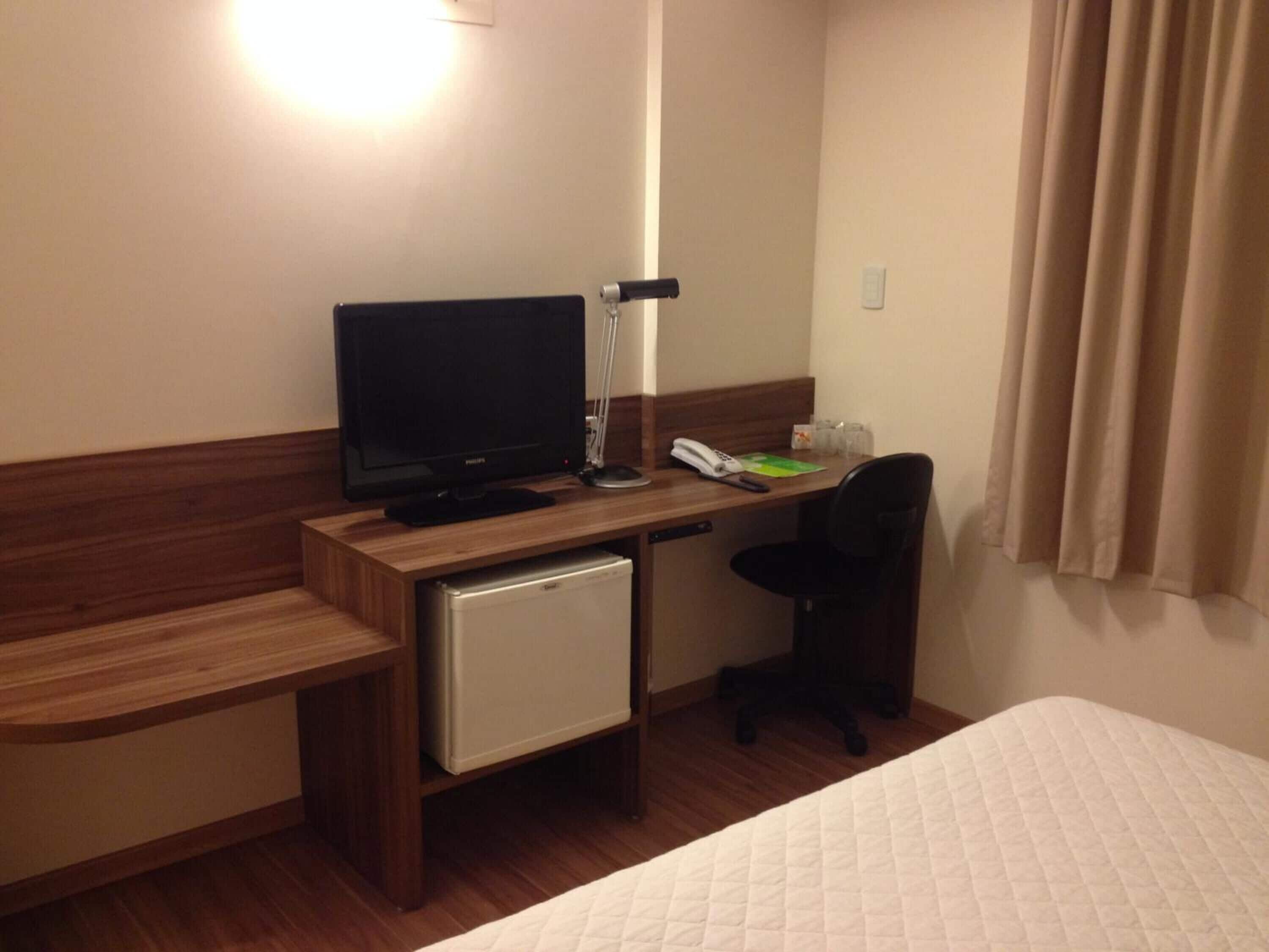 A hotel room with a wooden desk, a television, a small refrigerator, a telephone, and a bed with a quilted cover.