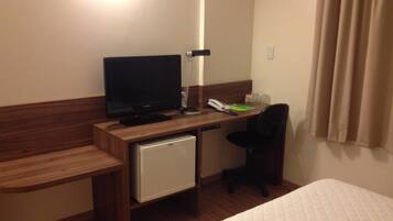 A hotel room with a wooden desk, a television, a small refrigerator, a telephone, and a bed with a quilted cover.