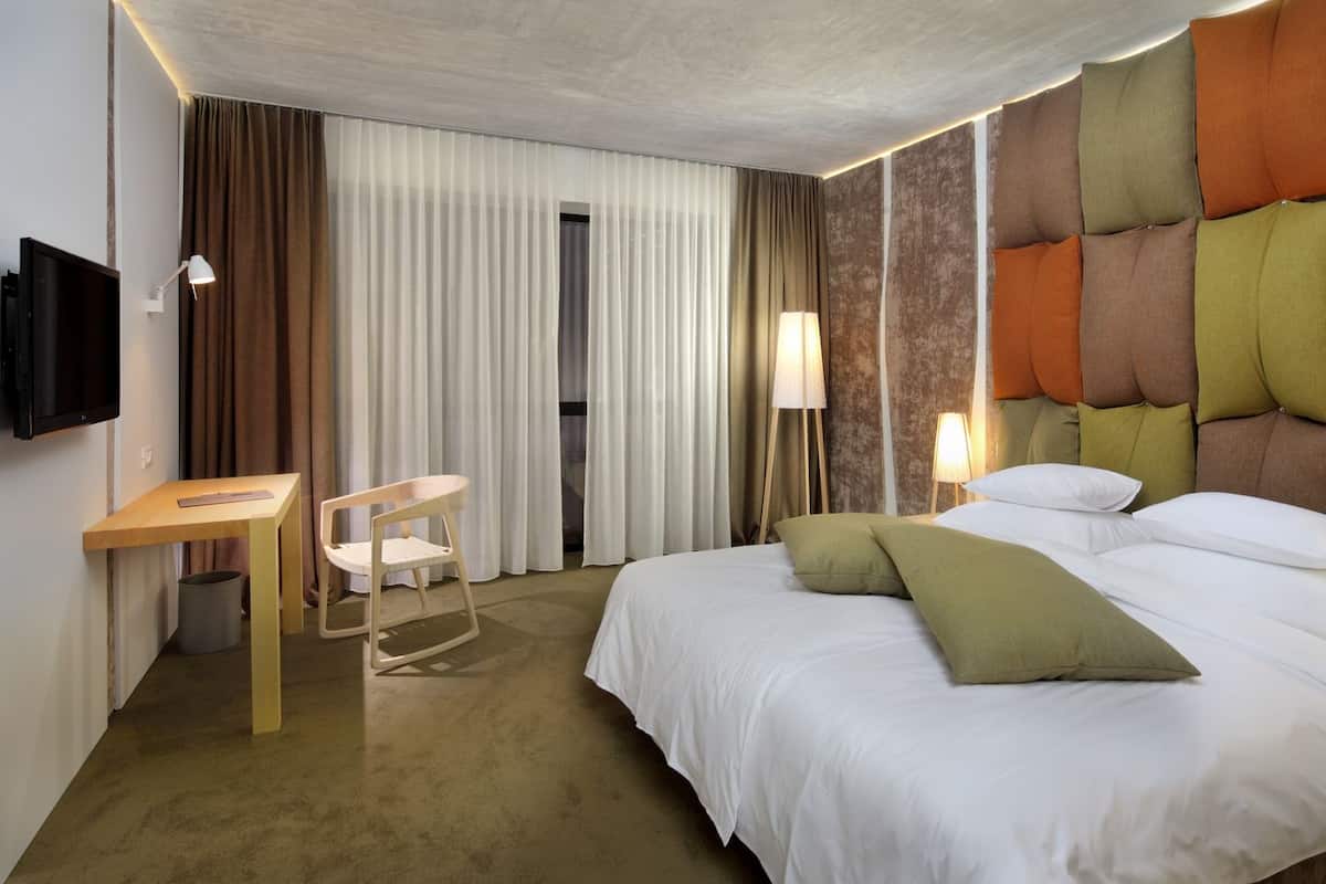 Habitación de hotel con cama, escritorio, silla, televisión y ventana con cortinas.