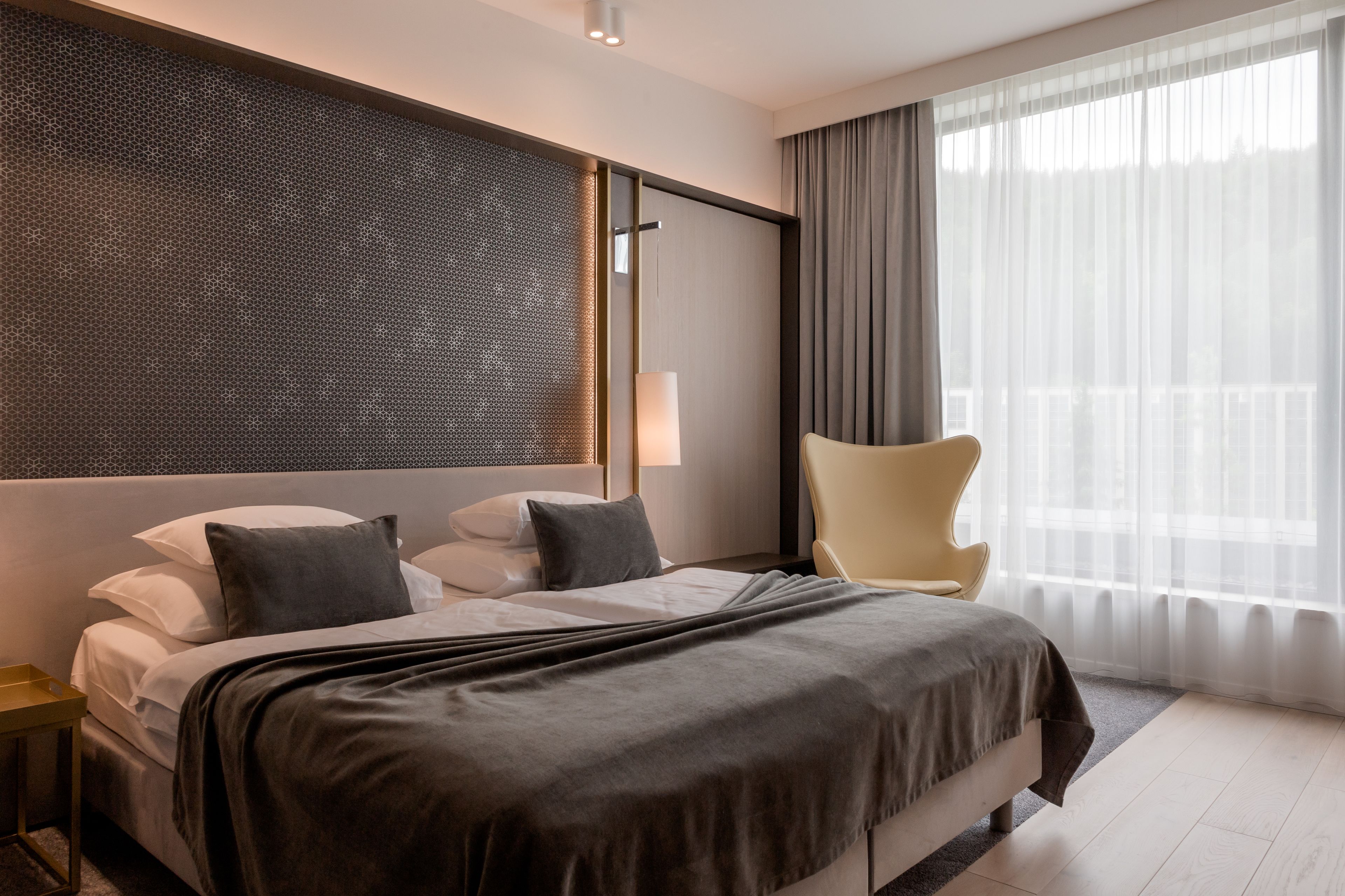 A modern hotel room with a large bed, a chair, a wall-mounted TV, and a large window with curtains.