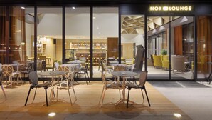 Un Nox Lounge con una decoración moderna, con mesas y sillas redondas, y una zona de bar visible a través del ventanal.