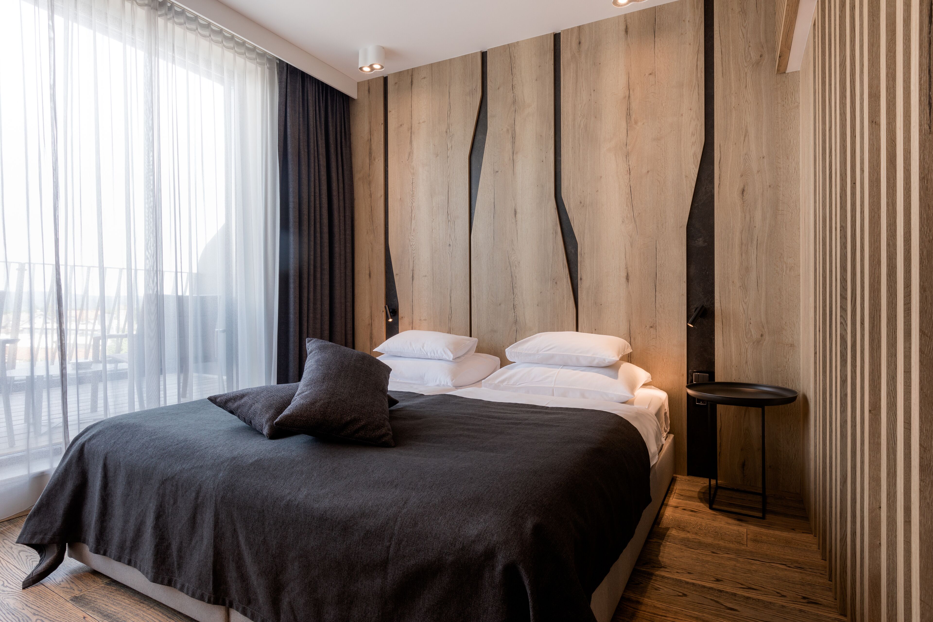 A modern bedroom with a large bed, a small side table, and a view of the outdoors through sheer curtains.