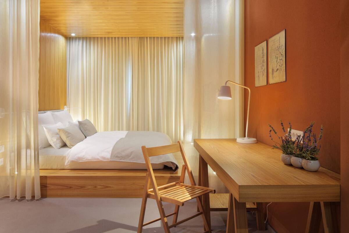 Un dormitorio moderno con cama, escritorio de madera, silla y una mesita con lámpara y plantas.