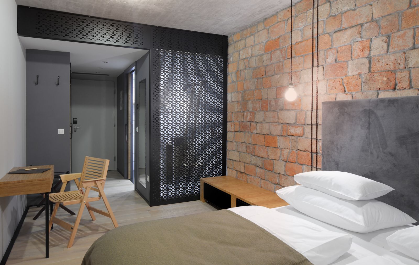 A modern bedroom with a brick wall, a bed with white bedding, a wooden bench, and a desk with a chair.