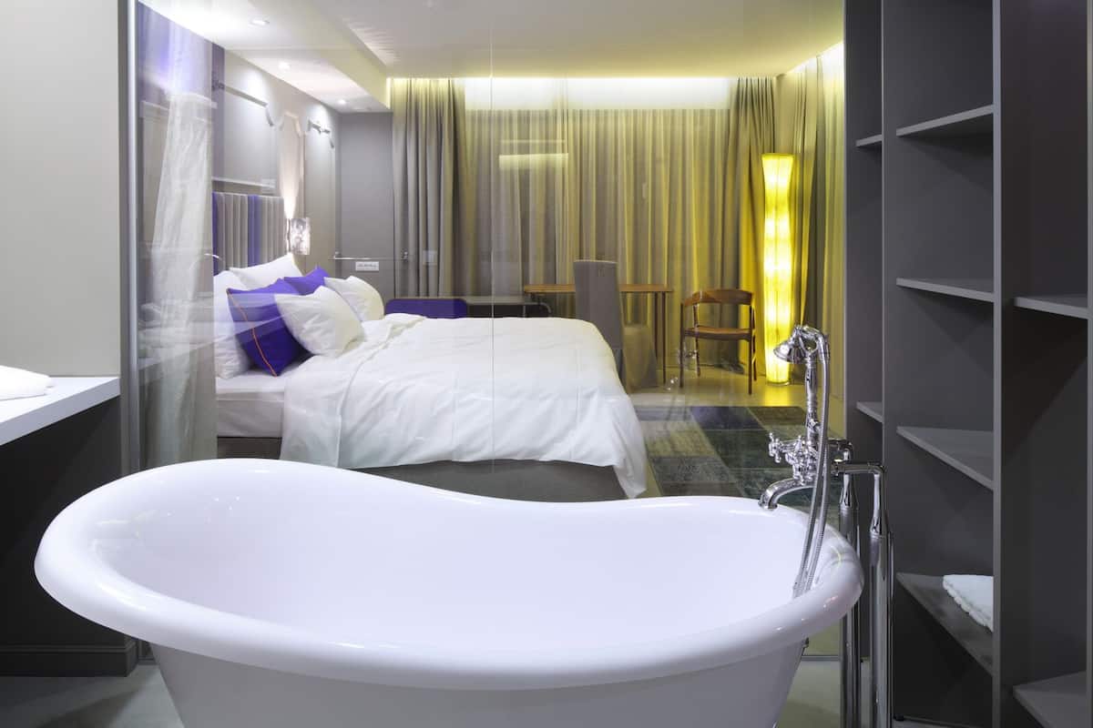 Una habitación de hotel moderna con una bañera grande, una cama bien hecha, una zona de estar con una silla y una mesa, y un espacio para la ducha.