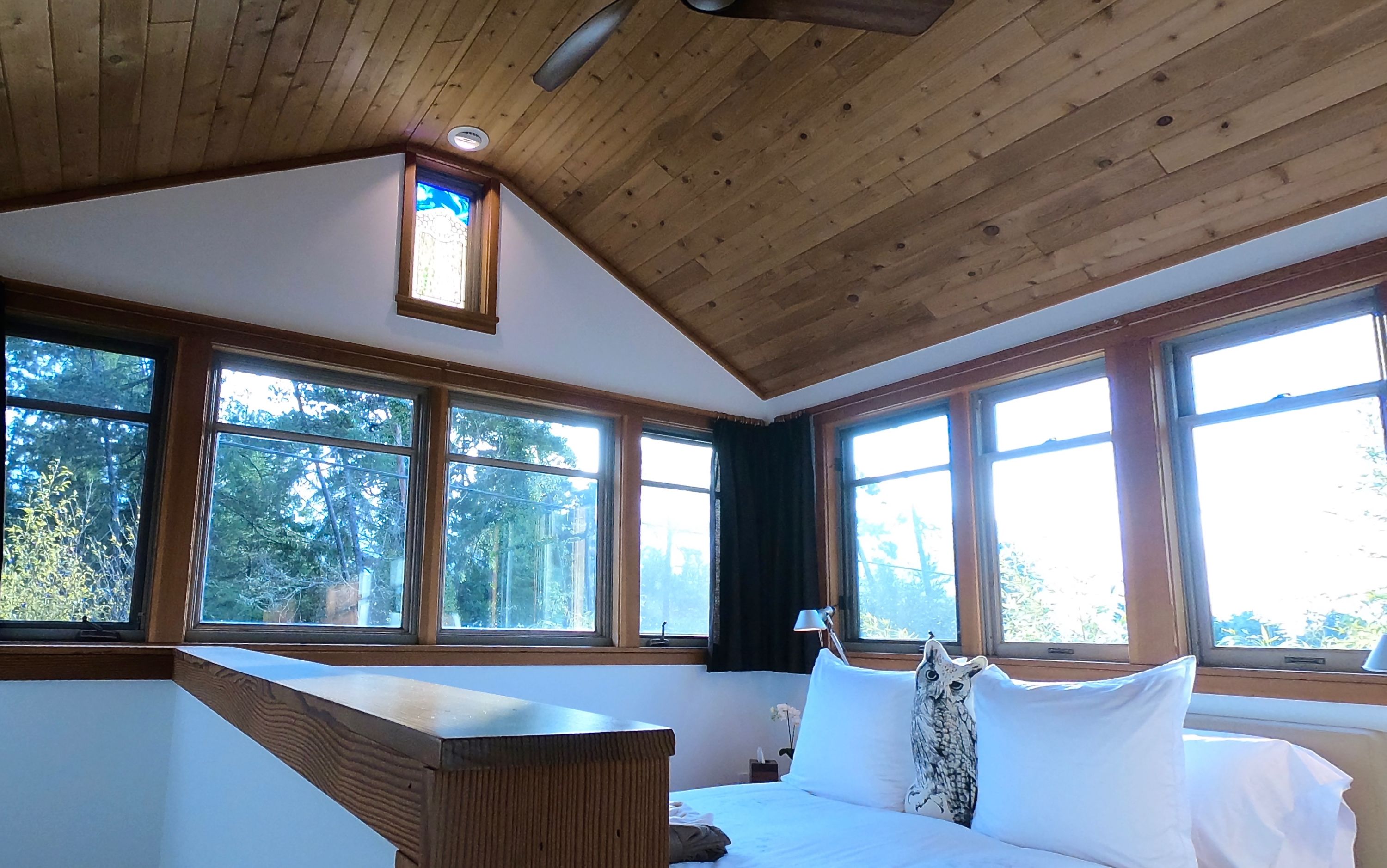 Una habitación con techo de madera, ventanas amplias, cama con ropa blanca y una almohada con forma de búho.